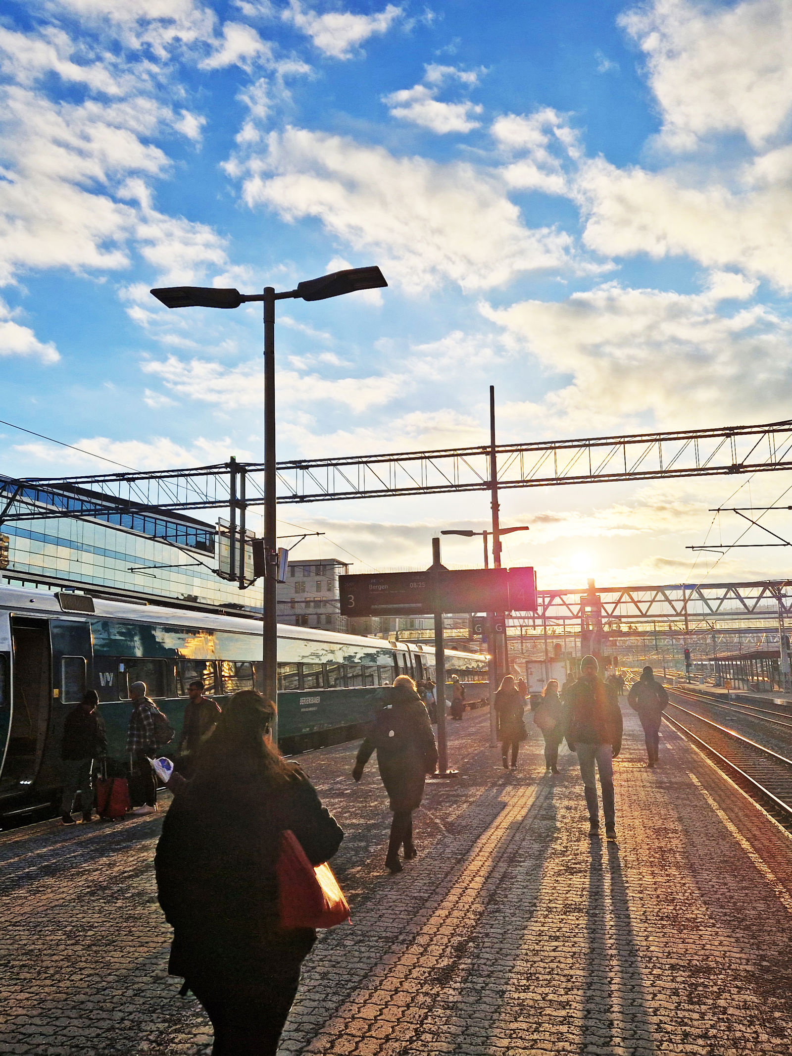 Lav sol over perrongene på Oslo S. Folk går på plattformen mot sola. Et tog står på ett av sporene.