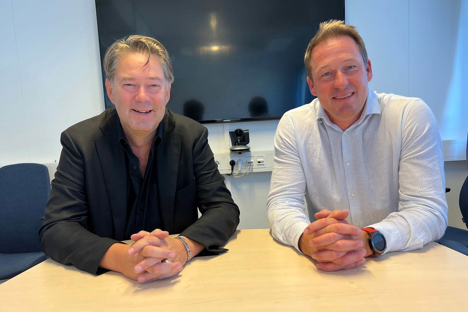 To menn sitter ved bord og smiler til kamera.