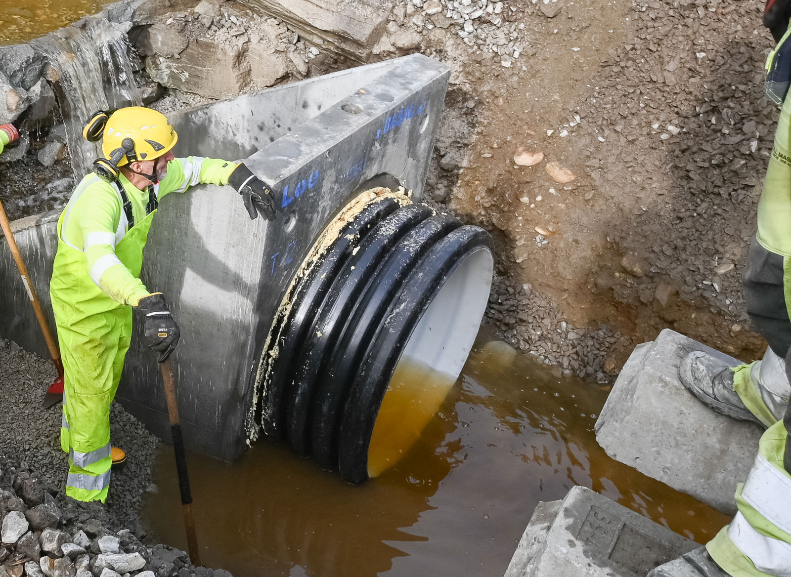 Tre menn i gule arbeidsklær installerer ny og større stikkrenne under jernbanespor-