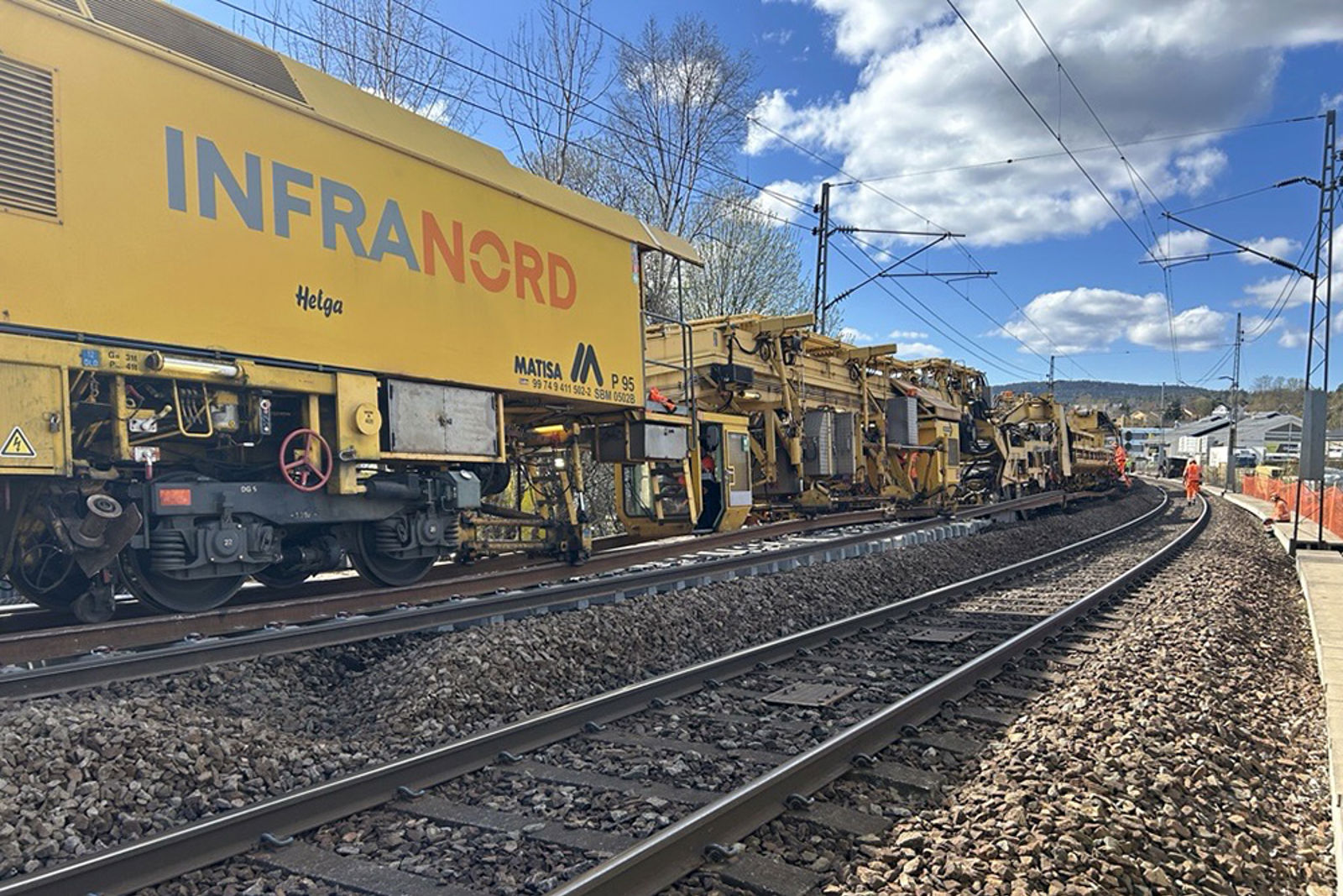 Det lange, gule arbeidstoget er i sving på det venstre av to spor. Arbeidere i bakgrunnen.