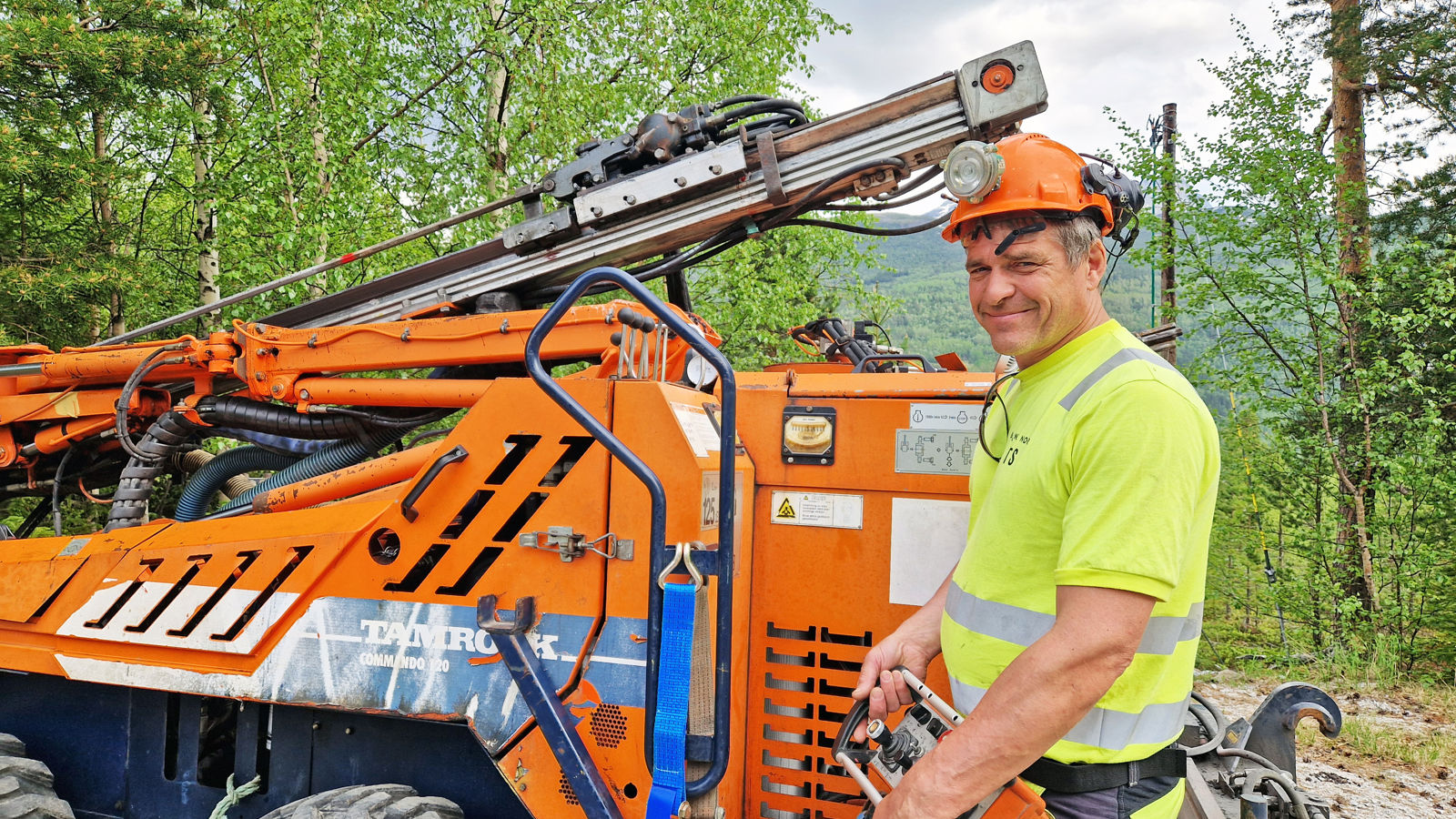 En mann i vernetøy står foran en oransje anleggsmaskin, som er en borerigg. 