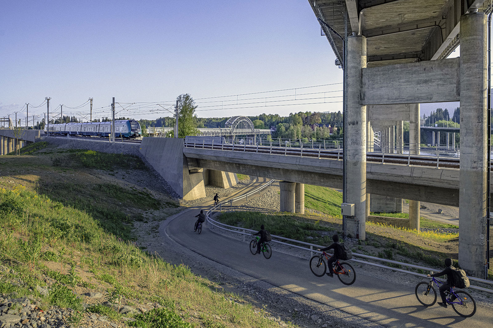 Syklister på turvegen under dobbeltsporet hvor et tog kjører. I høyden undersiden av vegbrua.