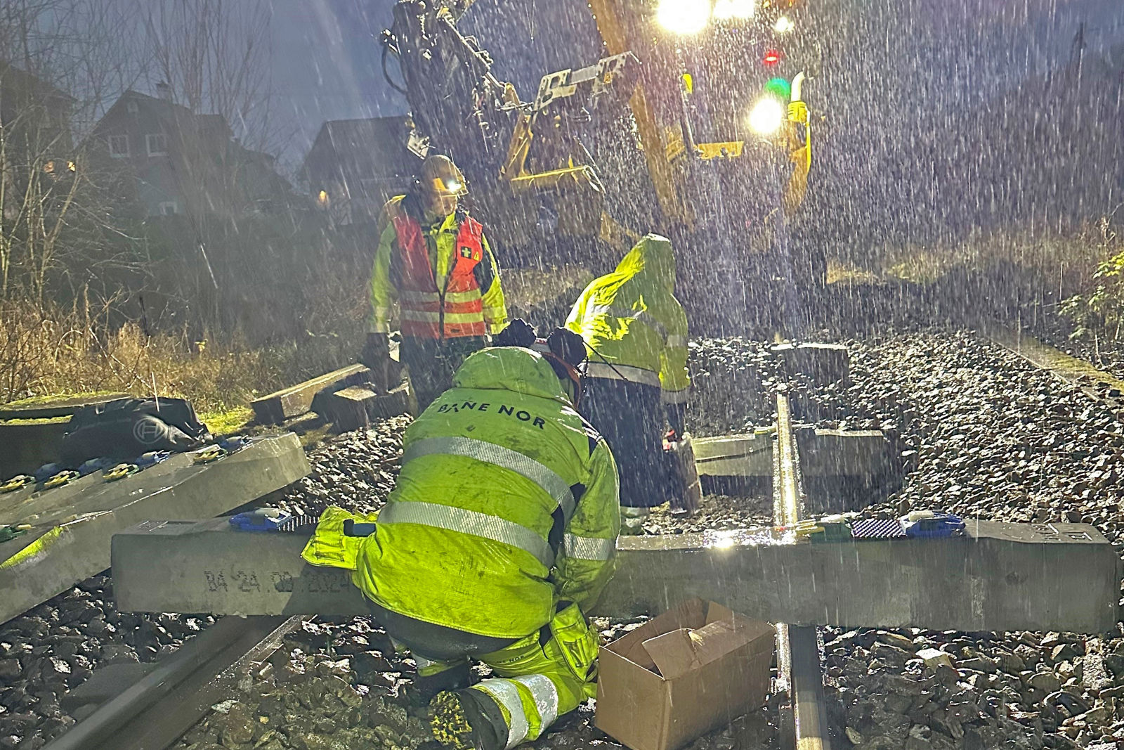 Tre personer i anleggsutstyr og en arbeidsmaskin i sving med svillebytte i mørke og styrtregn.