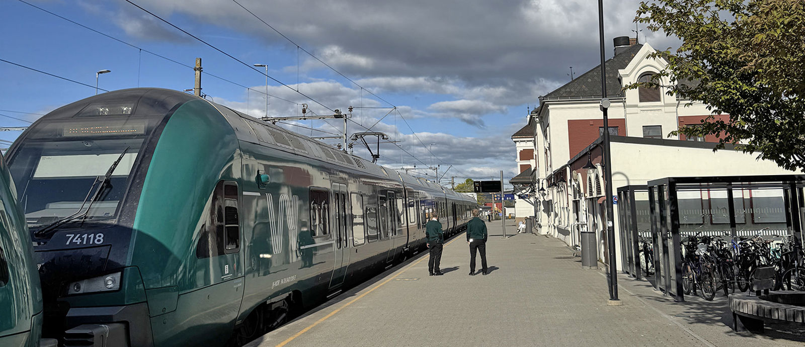 Grønt Vy-tog til venstre, personer på plattformen, stasjonsbygningen og sykkelparkering til høyre.