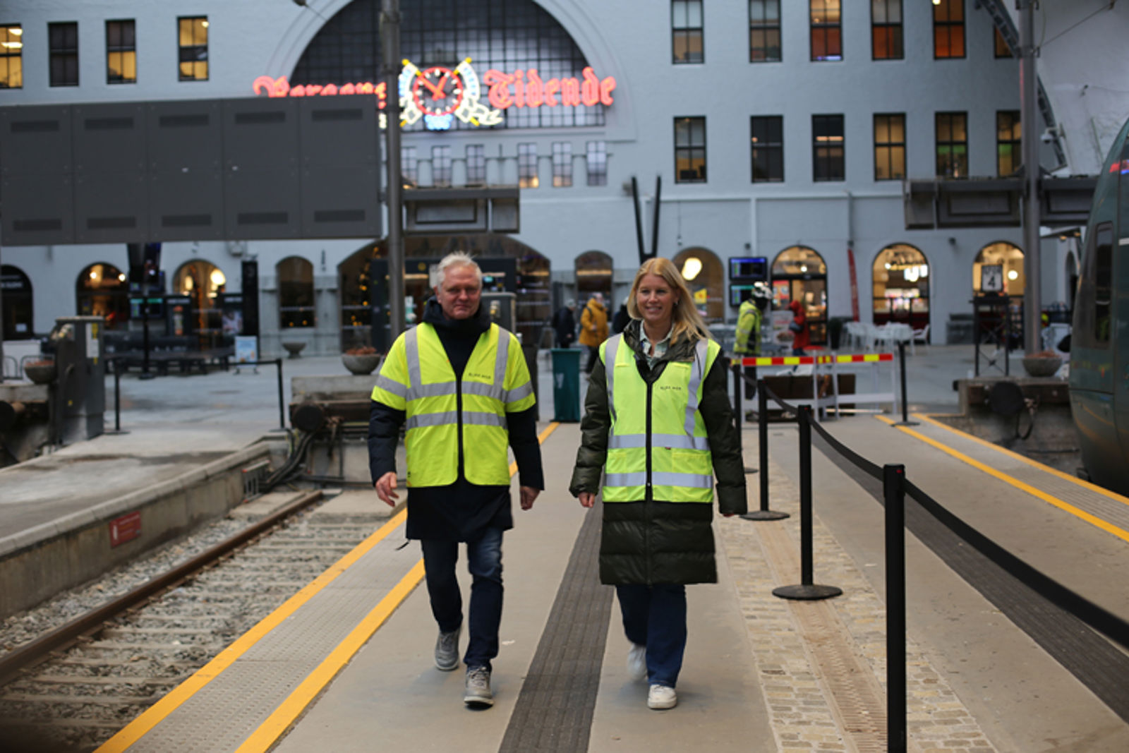Bildet viser to personer i gule vester som går på en jernbaneplattform