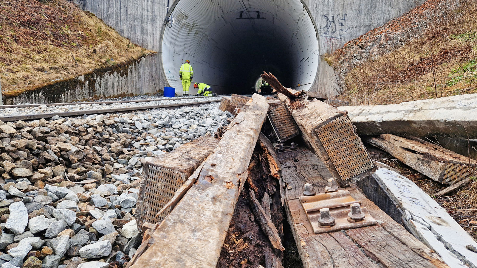 Noen gamle, råtne tresviller i forgrunn, jernbanespor som går inn i en tunnel i bakgrunn. 