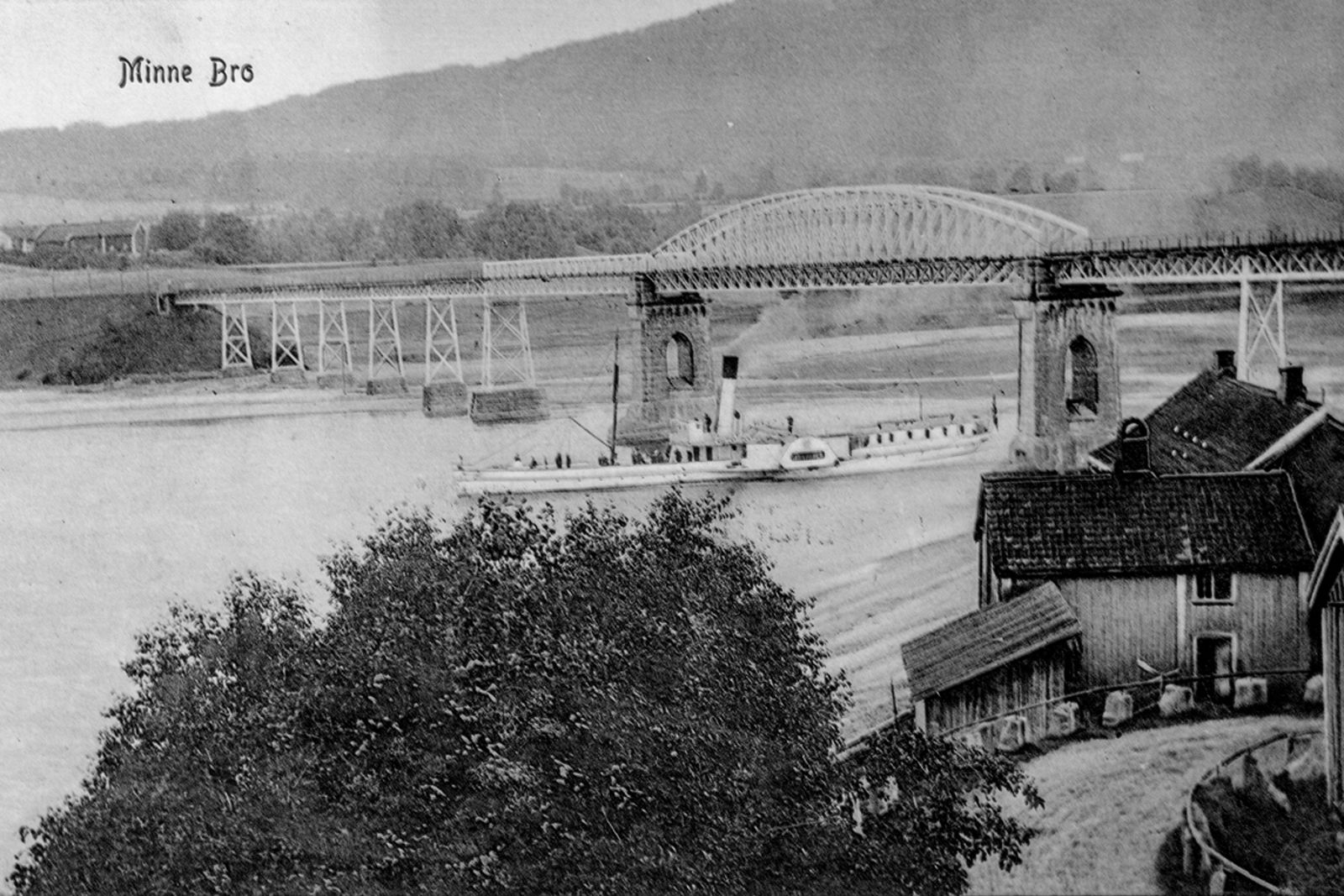 Trær og gårdsbygninger i forgrunnen, Skibladner på veg under brua.