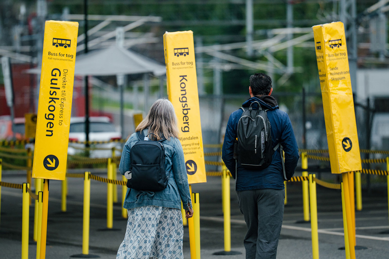 To personer med bagasje foran gule sperringer og skilt med buss for tog-destinasjoner.